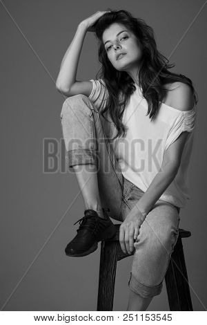 A Beautiful Brunette Girl In A White T-shirt And Jeans Sits On A High Chair, Lifting One Leg On The 