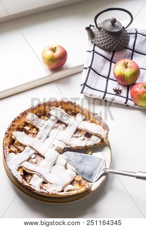 Homemade apple pie sprinkled with powdered sugar with a sliced piece  among apples, cinnamon sticks 