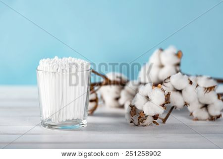 Cotton Swabs In Clear Glass Jar In Bathroom Setting With A Cotton Flowers On Pastel Blue Background.