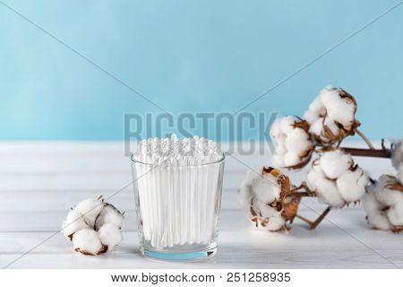 Cotton Swabs In Clear Glass Jar In Bathroom Setting With A Cotton Flowers On Pastel Blue Background.