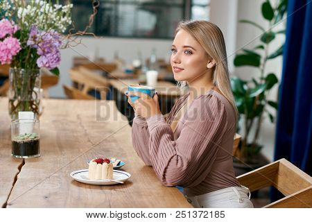 Beatiful Girl With Blonde Hair And Blue Eyes Drinking Coffee In Cafe. Keeping Cup, Wearing Beige Blo