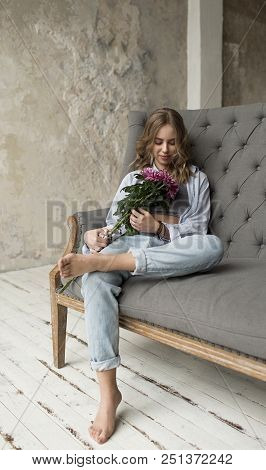 Blonde Beautiful Model With Curly Hair Sitting On The Grey Sofa. Girl Holding Pink Flowers.