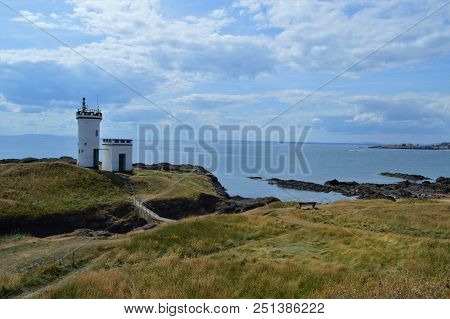 A White Ligthhouse On Small Hill Surrounded By The Sea And A Cloudy Sky In The Background