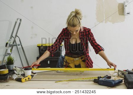 Beautiful blond young female putting spirit level on plywood sheet placed on carpenter workbench with tools and instruments