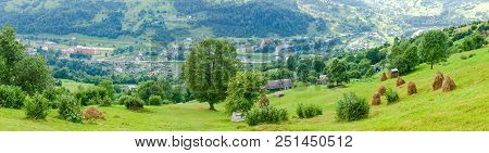 Wide Panorama Of The Mountain Village In Valley With Hayfield And Haystacks On Slope On A Foreground