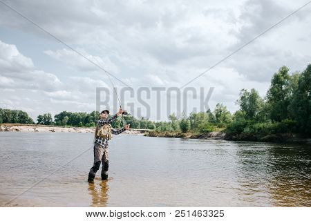 A Picture Of Man Standing In Water And Waving With Fly Rod. He Holds With Both Hands. Guy Is Looking