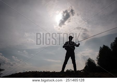 Dark Picture Of Fisherman Standsat The Edge Of Water. He Holds Fly Rod In Hads. He Is Fishing. Sun I