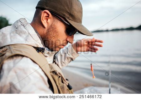 A Picture Of Man Sitting At The Edge Of Water And Looking Down. He Holds Spoon With Bait. Guy Is Pre