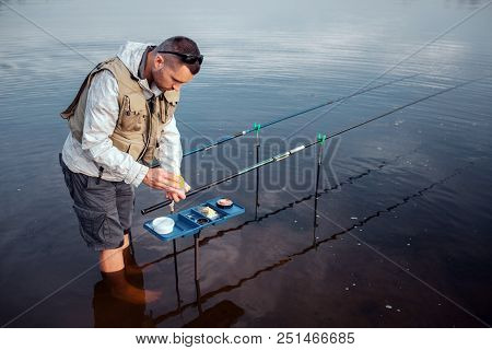 Young Fisherman Is Standing In Water Barefoot. He Is Leaning To Opened Plastic Box With Artificial B