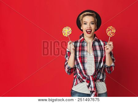 Surprised Girl With Two Striped Candies. Studio Portrait At Red Wall, Copy Space