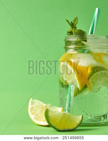 Mason glass jar on a green table with homemade sparkling lemonade with ice, slices of lime and lemon, green leaves of mint . Slices of citrus fruits on green background with copy space.