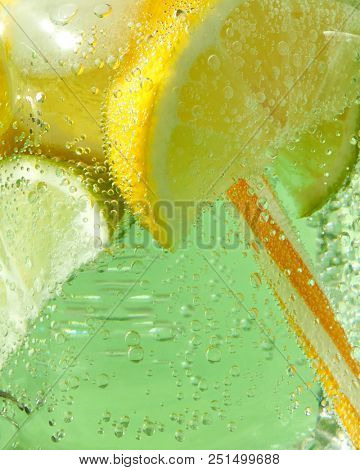 A macro shot of a glass with a cold refreshing lemonade with ice, pieces of lime, lemon and mint leaves, plastic straws on a green background. Bubbles of air in a glass jar.