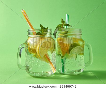 Cold natural lemonade handmade with bubbles of air. Citrus fruits slices of lemon and lime, ice, leaf of mint, water with plastic straws in the glass. Two glass jar on green background.