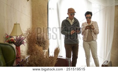 Asian Senior Couple Use Smart Phone Technology While Travel With Suitcases In Luxury Interior