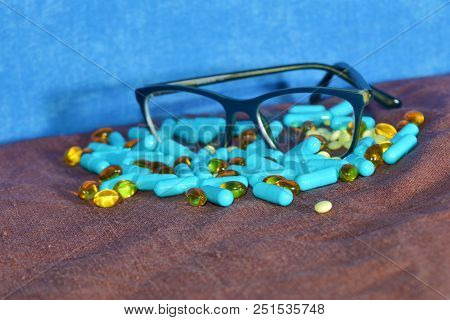 Blue Capsules And Different Pills Are Scattered On The Brown Surface Of The Rowades With Glasses On 