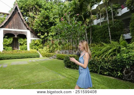 Young Nice Woman Using Smartphone And Standing With Resort House In Background.
