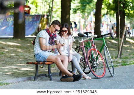 Young Couple In Love With Bicycle Sitting On Beanch And Looking Something In The Smartphone In The S