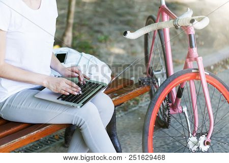 Pretty Girl With Bicycle Sitting On Beanch And Using Laptop In The Sunny Summer Park