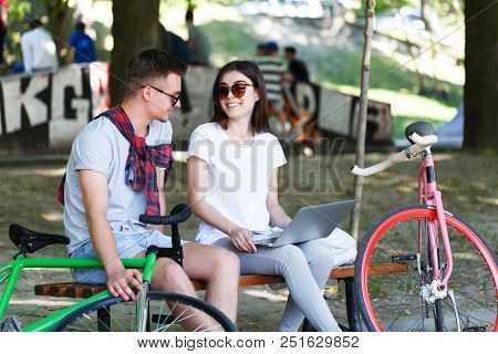 Lovely Couple With Green And Pink Bicycle Sitting On Beanch And Using Laptop In The Sunny Summer Par