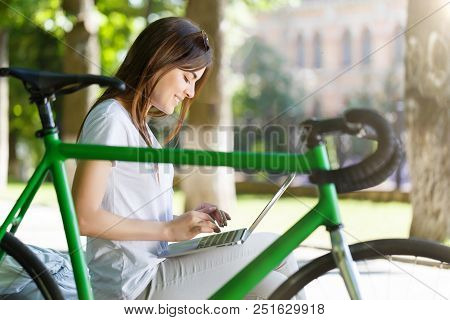 Young Cute Girl With Green Bicycle Sitting On Beanch And Working With Laptop In The Sunny Summer Par