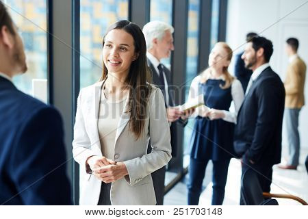 Smiling pretty young business lady in jacket talking to colleague and discussing business forum topics during break