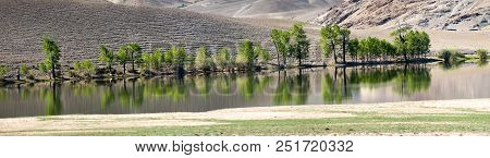 Green Trees And Bushes Are Reflected In The Water Of The Lake, Behind The Mountains Stretch, A Panor