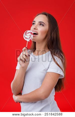 A Positive Girl With Dark Long Hair Is Licking A Sugar Candy, Posing On The Red Background.