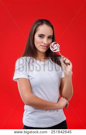 A Positive Girl With Dark Long Hair Is Licking A Sugar Candy, Posing On The Red Background.