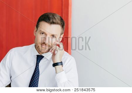 Indoor Shot Of Self Confident Businessman In White Shirt With Tie, Keeps Hands Near Face, Waits For 