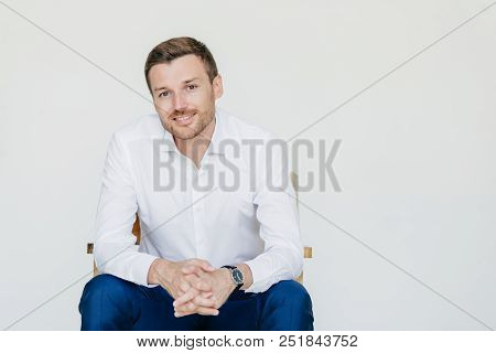 Studio Shot Of Elegant Cheerful Male Entrepreneur In Formal Clothes, Sits On Chair, Poses At Camera,