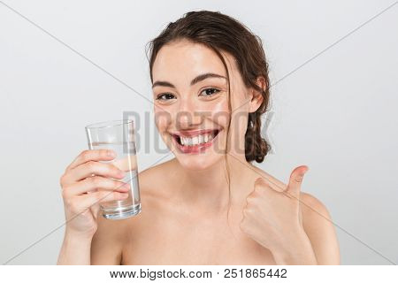 Beauty portrait of a smiling young topless woman with make-up holding glass of water and showing thumbs up isolated over gray background