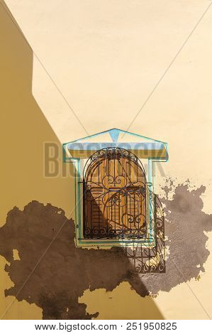 Window With An Arch, Closed Wooden Shutter And A Lattice. Green-blue Frame. Yellow Wall, Partly Repa
