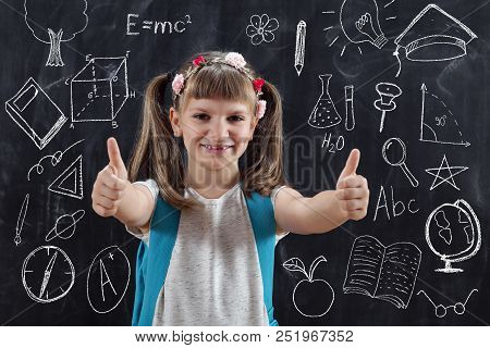 Schoolgirl Wearing Backpack, Standing In Front Of Blackboard And Showing Thumbs Up On Her First Day 