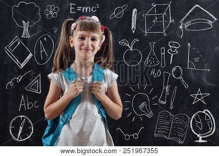 Schoolgirl Wearing Backpack, Standing In Front Of A Blackboard On Her First Day Of School