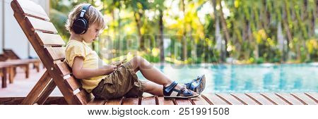 A Cute Little Boy Is Using A Smartphone And Headphones Lying On A Deckchair By The Pool. Primary Edu