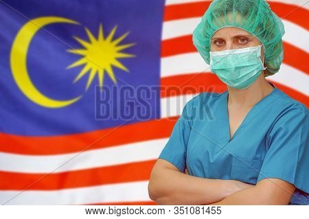 Female Surgeon In Mask And Hat Looks At The Camera On The Background Of The Malaysia Flag. Health Ca