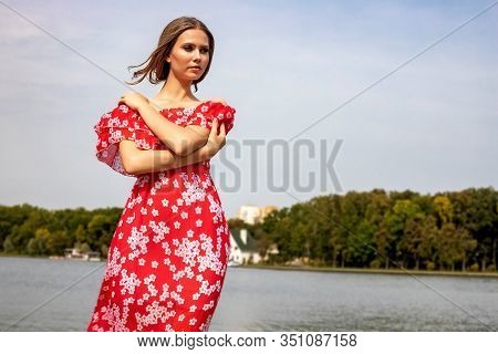 Portrait Of A Beautiful Young Brown-haired Girl With Long Hair In A Red Dress On A Lake Background.