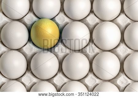 Identical White Eggs In A Box And One Yellow Gold Easter Egg Stand Out From The General Background. 
