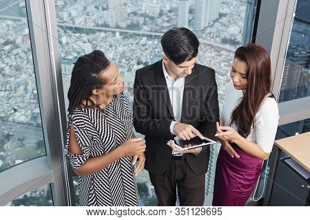 Positive Multi-ethnic Coworkers Standing At Big Office Wondow And Discussing Information On Tablet C