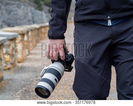 Hand Of A Man Holding A Camera With A Big Zoom Objective Outdoors In The Middle Of A Path Surrounded