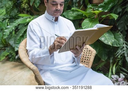 Saudi Muslim Man Dress Hijab Sitting And Writing On Textbook,freelance Business Concept.