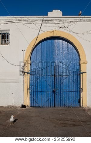 White Facade Of A Building With Blue Wooden Door With An Arch. Yellow Frame. Bright Blue Sky Matchin