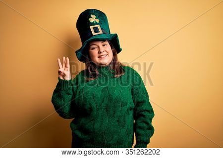 Young beautiful plus size woman wearing green hat with clover celebrating saint patricks day showing and pointing up with fingers number three while smiling confident and happy.
