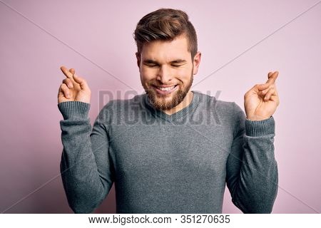 Young handsome blond man with beard and blue eyes wearing casual sweater gesturing finger crossed smiling with hope and eyes closed. Luck and superstitious concept.