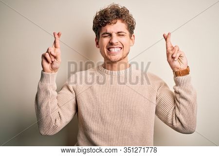 Young blond handsome man with curly hair wearing casual sweater over white background gesturing finger crossed smiling with hope and eyes closed. Luck and superstitious concept.