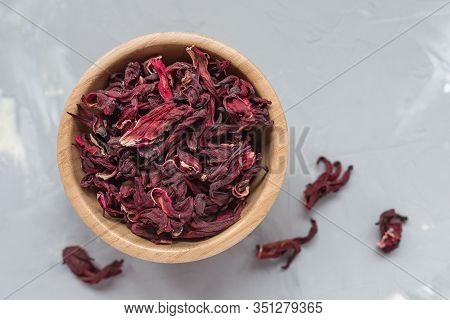 Dry Tea Of Red Hibiscus Petals, Also Known As Karkade Or Roselle Fruit, In Wooden Bowl On Gray Backg