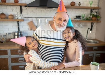 Two Dark-skinned Girls In Bday Hats Hugging Their Granddad