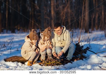 Family With Two Children, Daughters, And Dog Sitting By The Fire In Winter Snowy Forest And Roast On