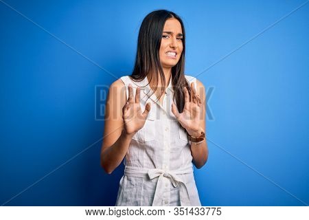 Young beautiful brunette woman wearing casual dress over isolated blue background disgusted expression, displeased and fearful doing disgust face because aversion reaction. With hands raised