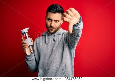 Young handsome sportsman drinking bottle of water to refeshment over red background with angry face, negative sign showing dislike with thumbs down, rejection concept